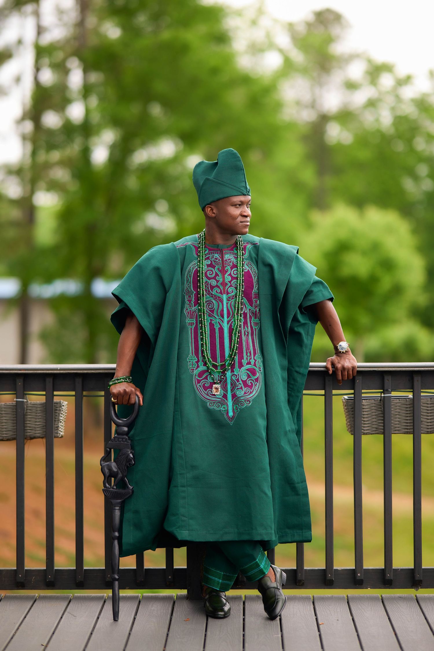 Man in green traditional African agbada outfit and matching cap poses on a deck with lush trees behind.