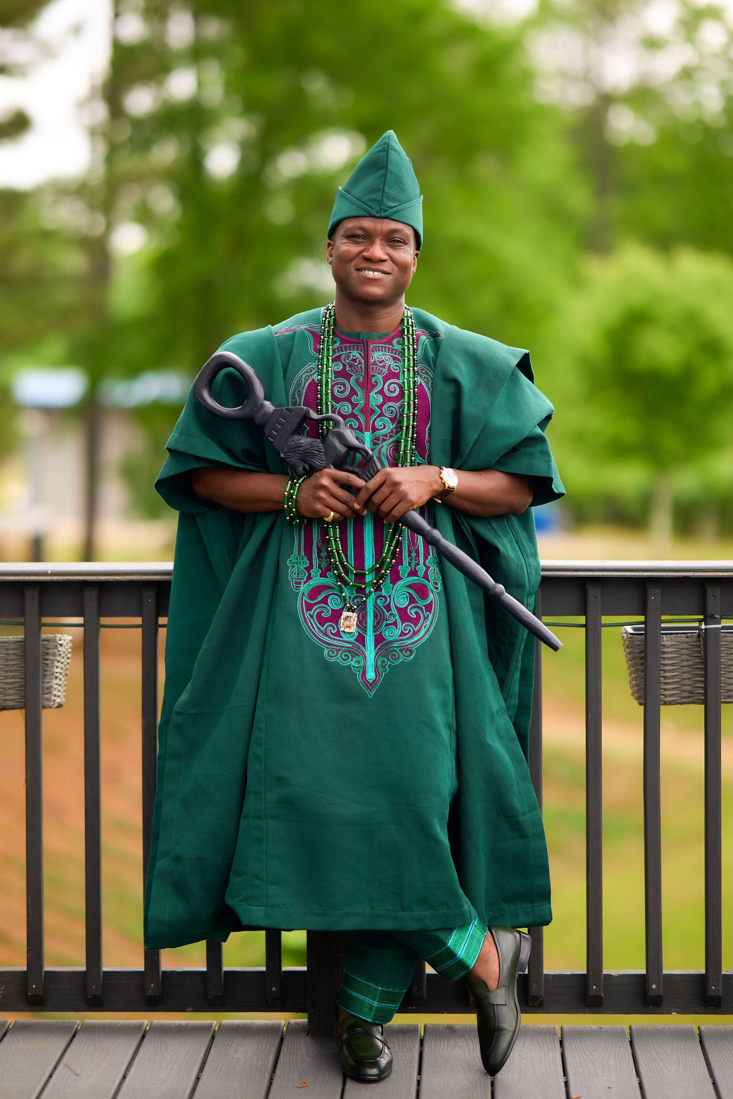 Man in green Nigerian agbada traditional attire with matching hat holding a ceremonial staff outdoors.