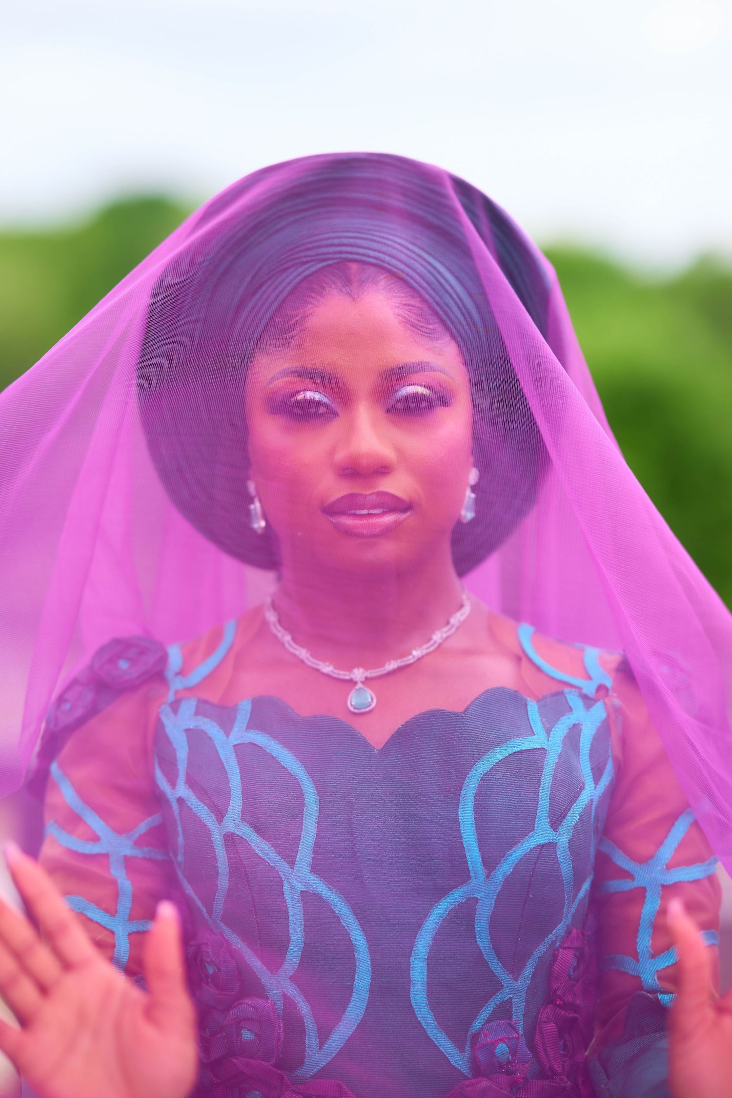 A Black woman in a blue embroidered gown and purple veil with matching eyeshadow poses gracefully outdoors.