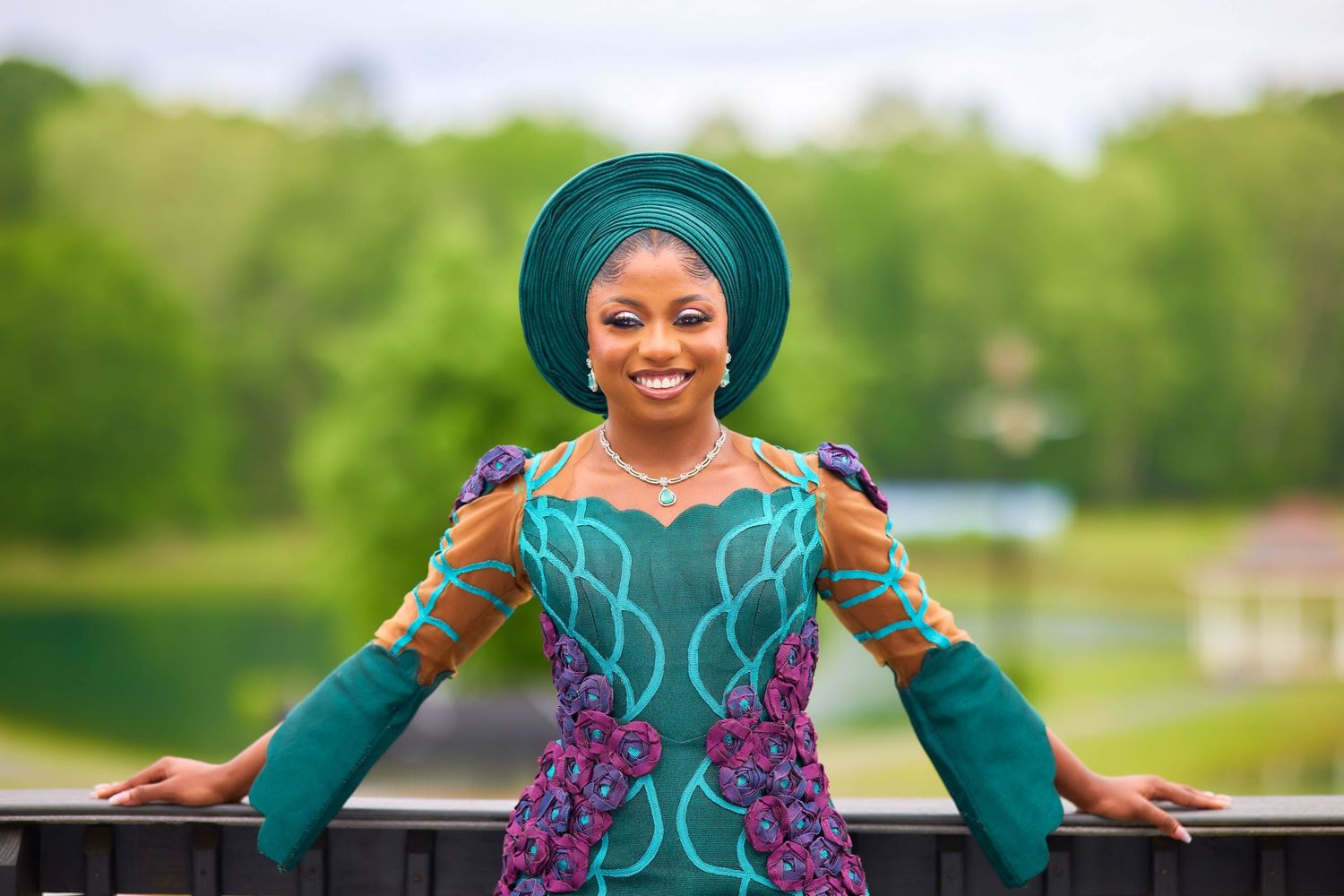 Woman in vibrant teal and purple African traditional attire with matching gele headwrap smiling outdoors.