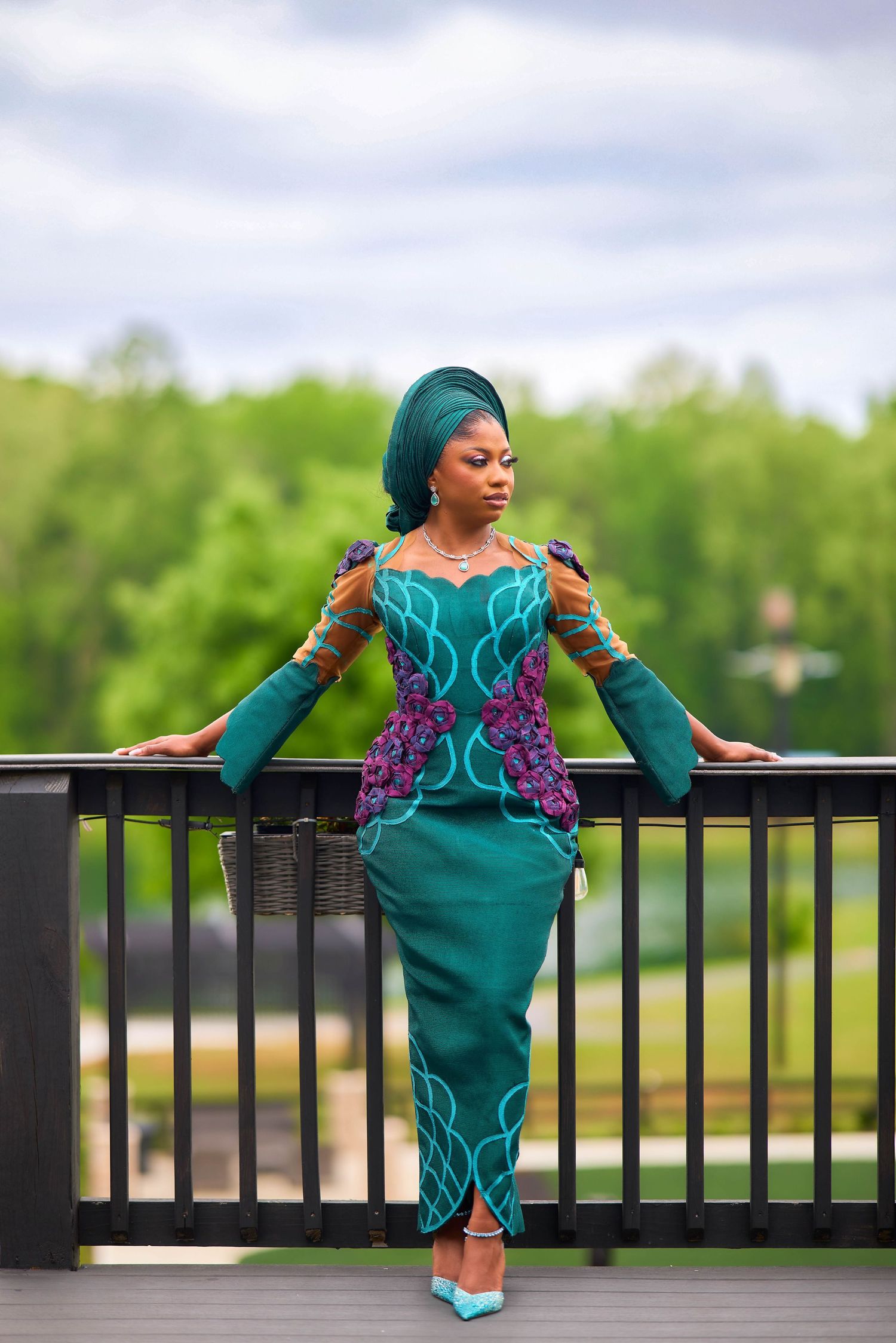 Woman in teal African dress and gele headwrap poses gracefully on outdoor balcony with lush green trees behind her.