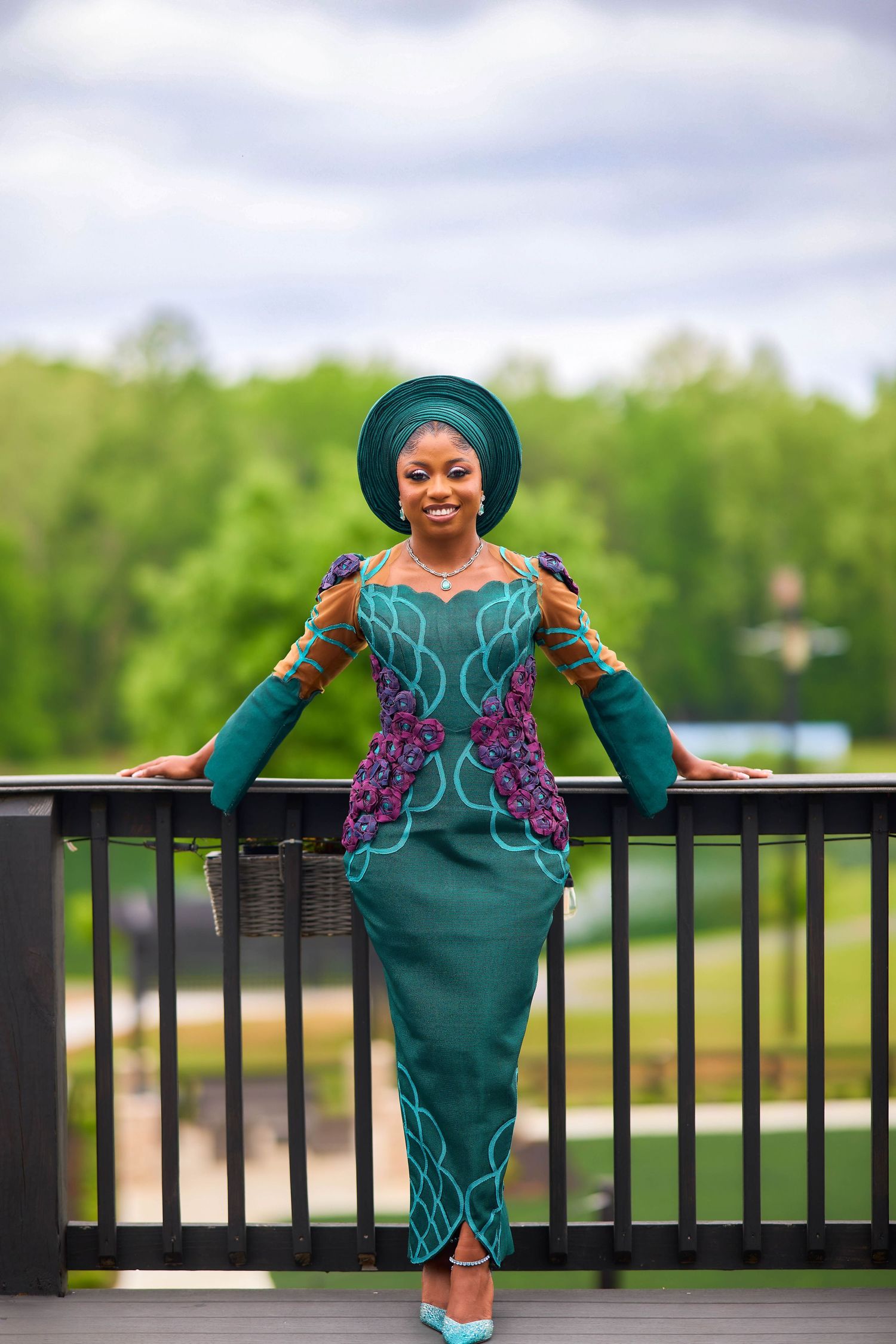 Woman in elegant teal African traditional attire with purple floral embroidery and gele headwrap posing on outdoor deck.