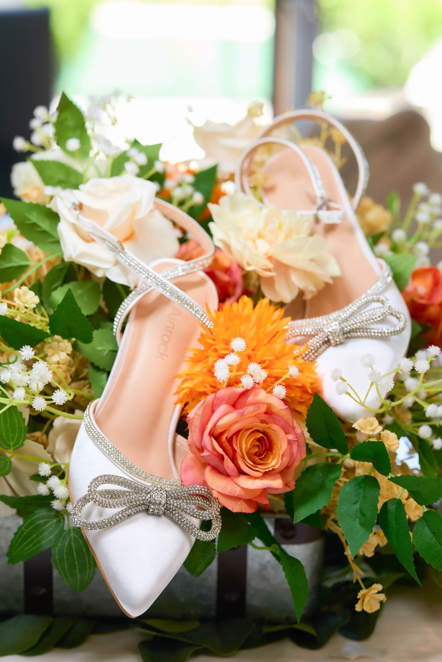 White satin bridal heels with crystal bow details resting on a colorful floral arrangement with roses and greenery.
