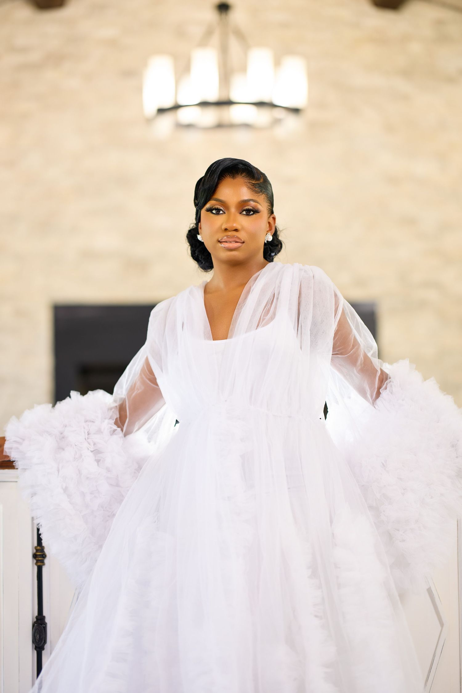 Young Black bride in elegant white tulle ballgown with sheer sleeves poses in a chandelier-lit venue.
