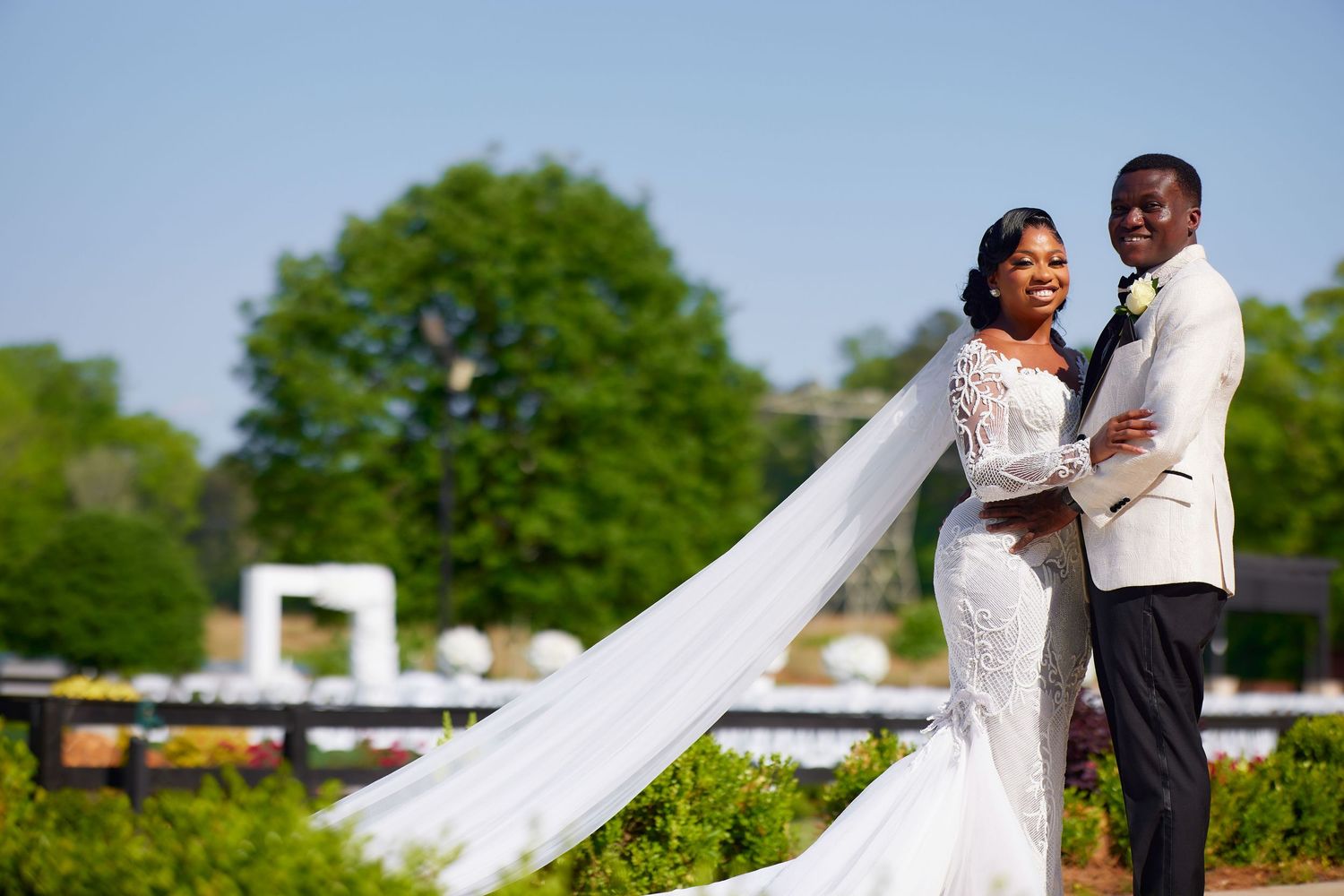Elegant bride in lace gown with flowing veil stands beside groom in white tuxedo outdoors on sunny wedding day.