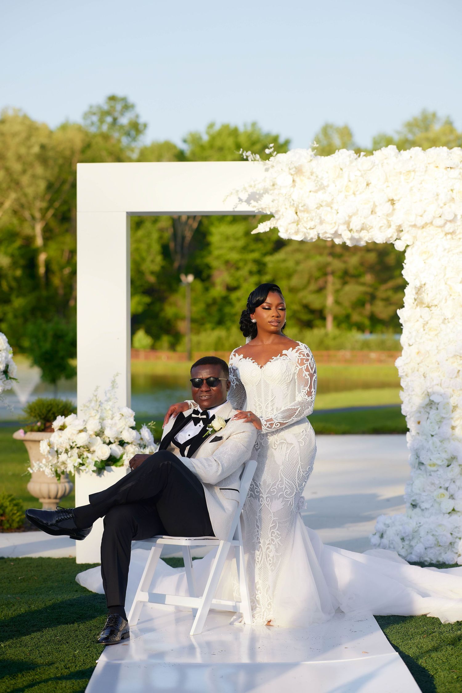 Elegant Black couple poses at outdoor wedding ceremony with white floral arch and lush greenery backdrop.