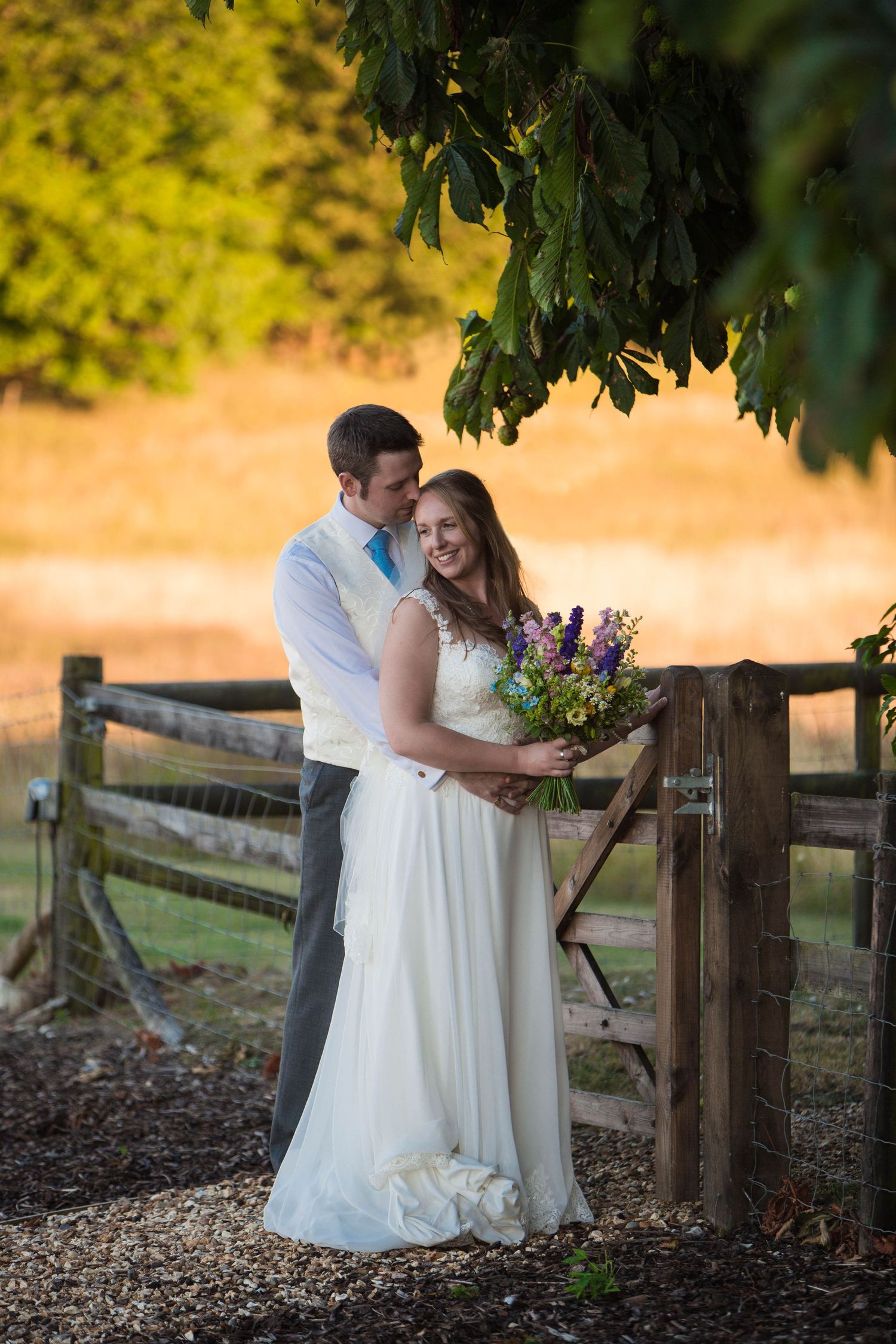 Bride and groom embrace by rustic wooden fence, she holds purple bouquet in white gown, golden sunset backdrop.