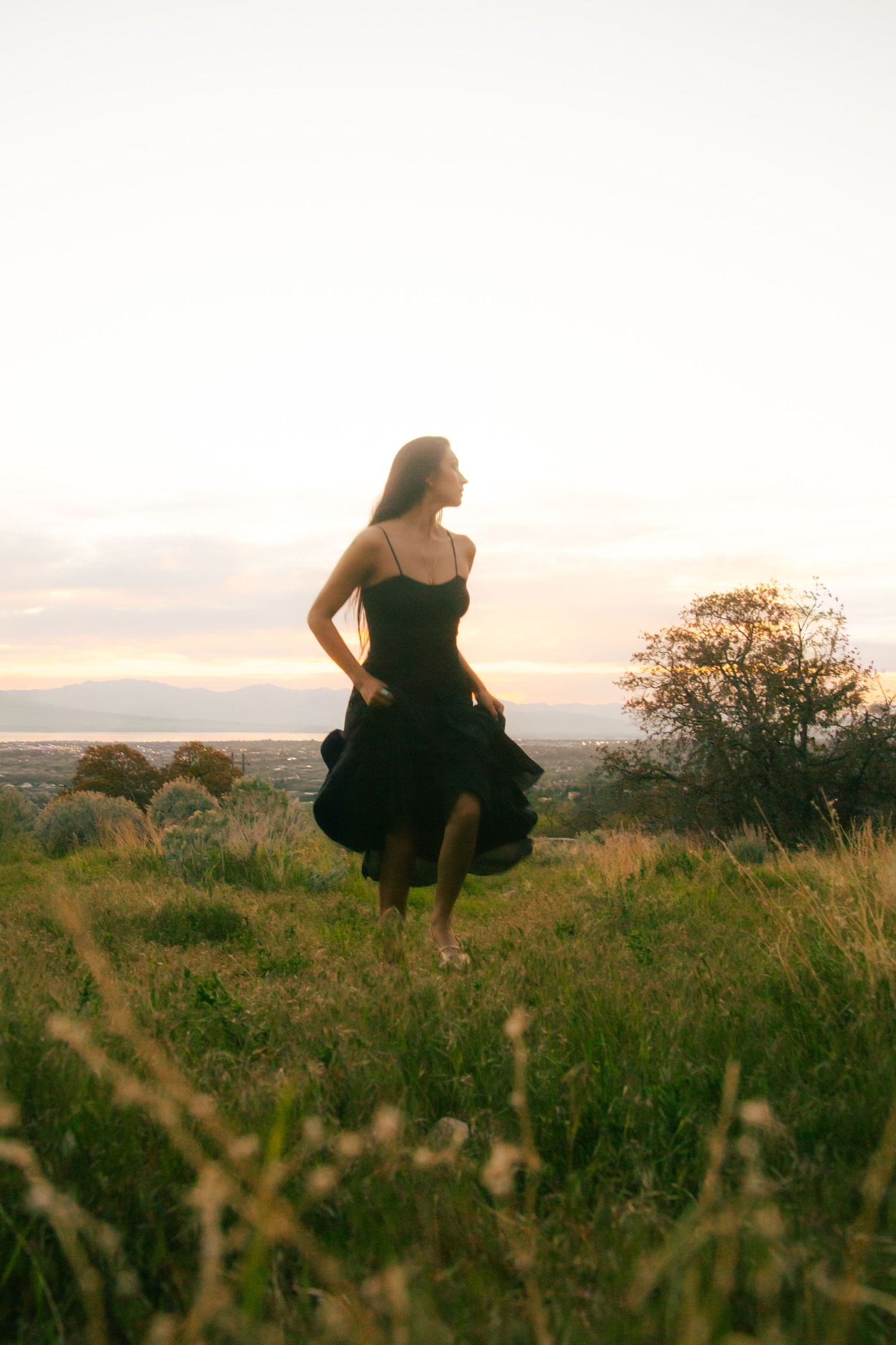 Woman in black dress walking through green meadow at golden sunset with scenic valley view.