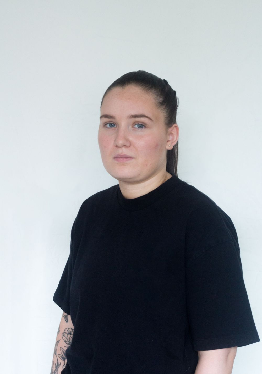 Young woman with dark ponytail in black oversized t-shirt with tattoo on arm against white background.