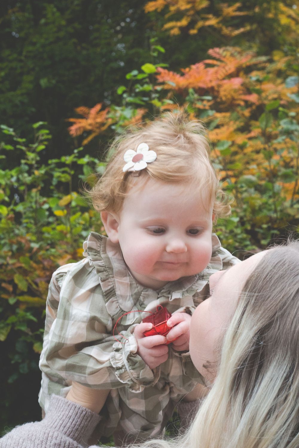 Mother holding a cute baby girl with a flower clip in her hair outdoors with autumn foliage behind.