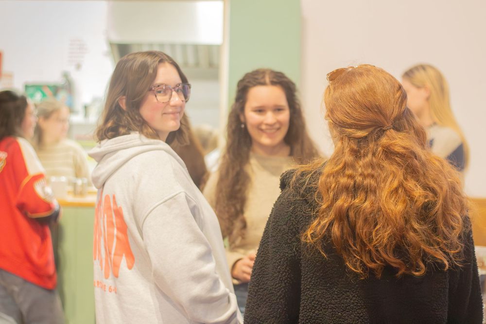 Three young women chat together in a casual indoor gathering, one wearing glasses and a gray hoodie.