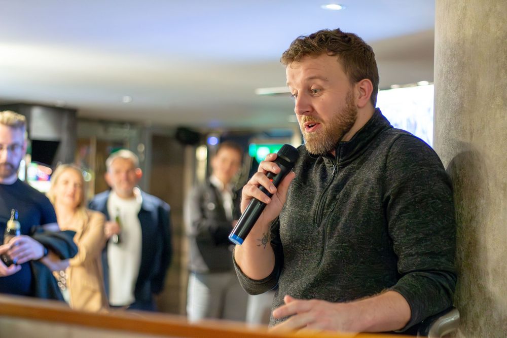 Man in grey sweater speaking into a microphone at a social gathering or event.