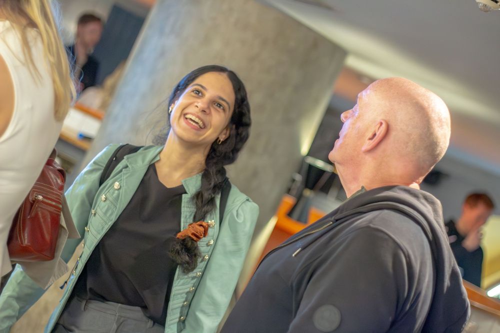 A woman in a green jacket smiles warmly while conversing with a bald man in an indoor social setting.