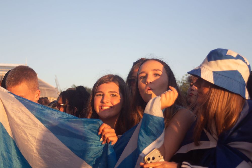Three young female festival fans wrapped in Scottish flags smiling at outdoor music event in golden evening light.