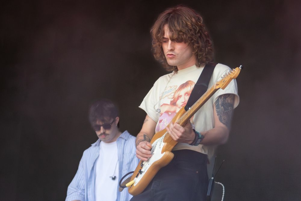 Young curly-haired guitarist playing electric Fender Stratocaster on smoky outdoor festival stage.