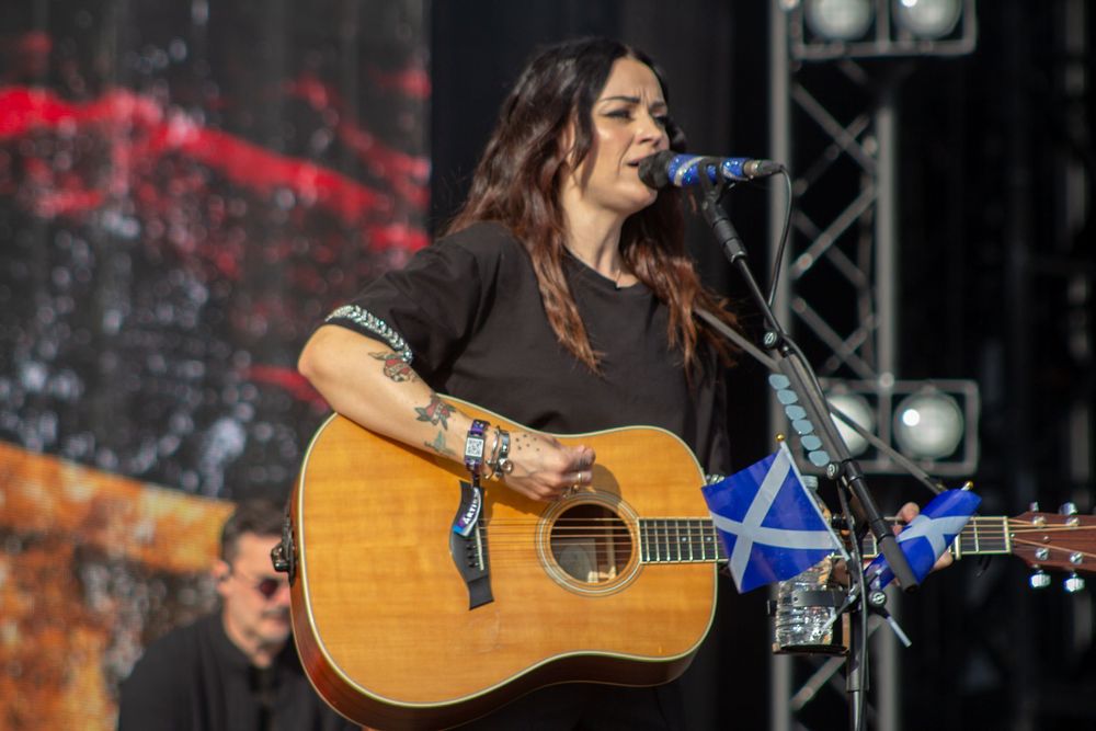 Dark-haired female musician playing acoustic guitar with Scottish flag decoration while singing at outdoor festival.