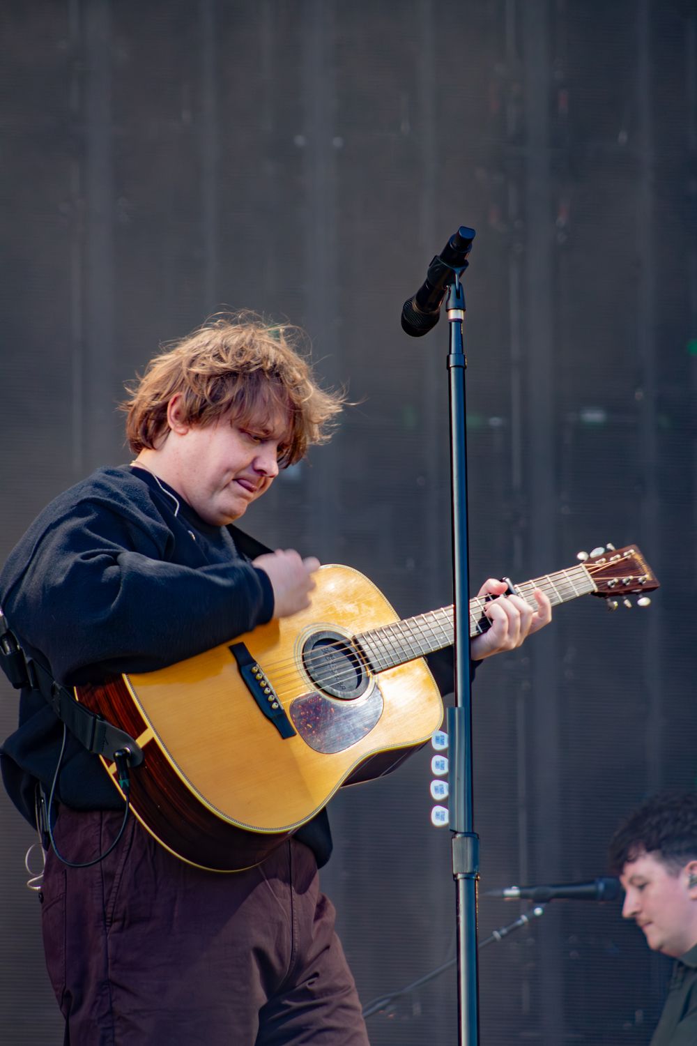 Young male guitarist with tousled hair playing acoustic guitar on an outdoor concert stage.