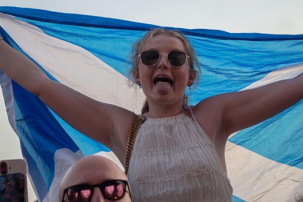 Enthusiastic woman holding a Scottish flag with arms outstretched, wearing sunglasses at an outdoor event.