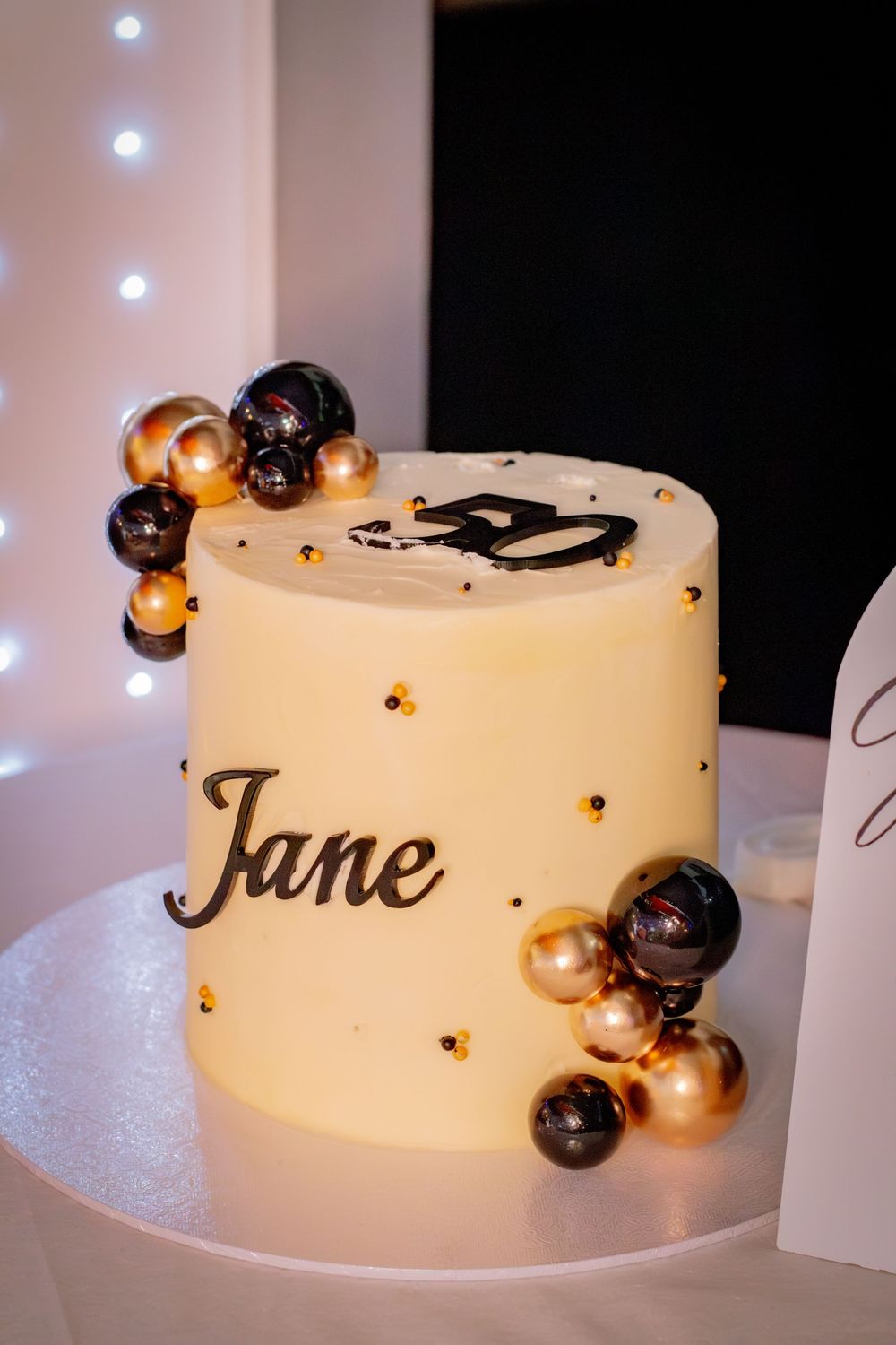 Elegant white birthday cake decorated with gold and black spheres reading Jane for 50th birthday.