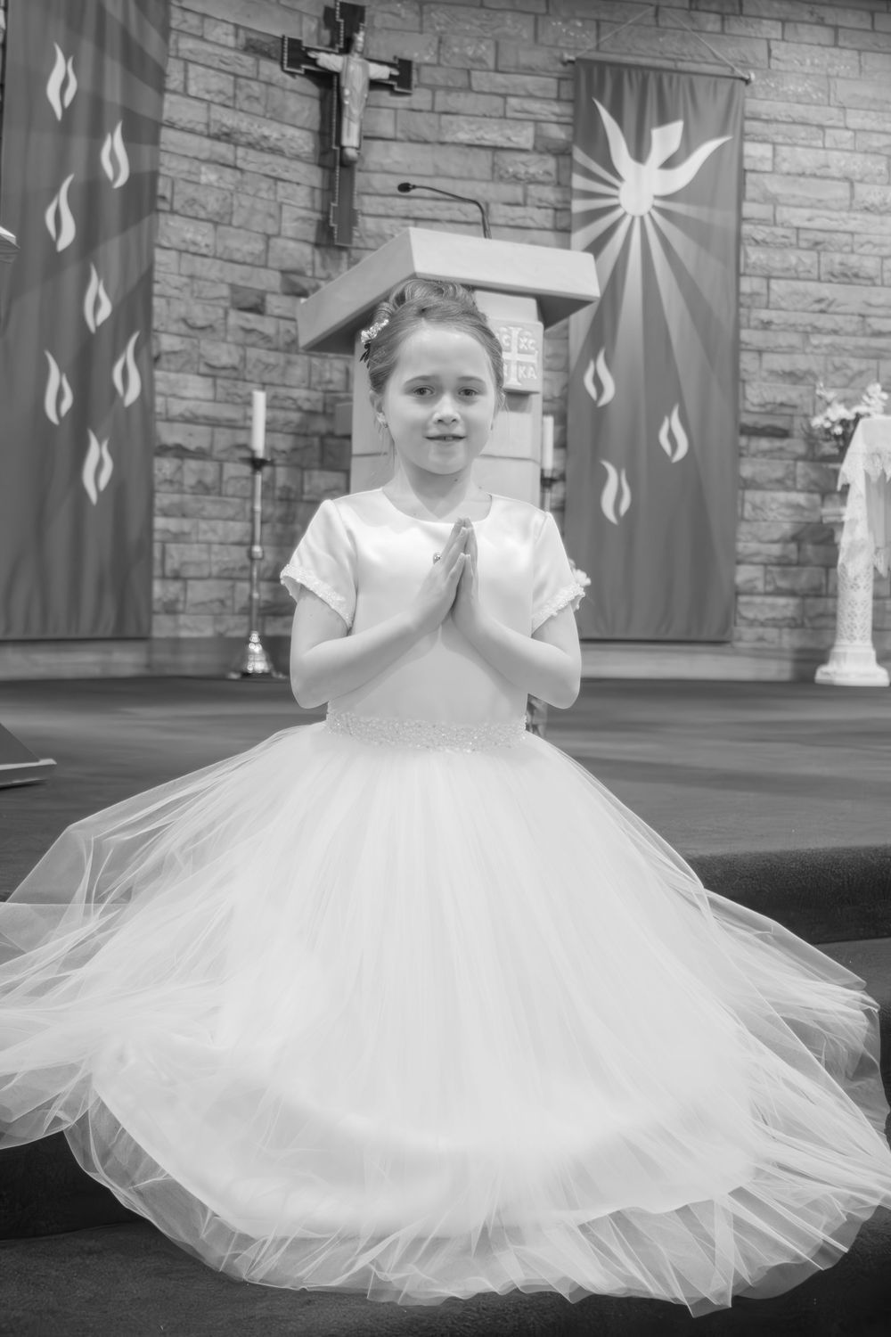 Girl in white communion dress stands with hands in prayer at church altar in black and white photo.