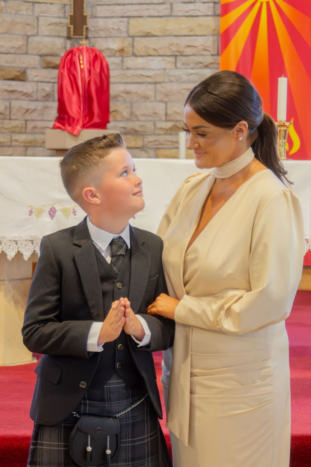 Boy in suit and kilt with hands in prayer poses with woman in cream dress at church altar.