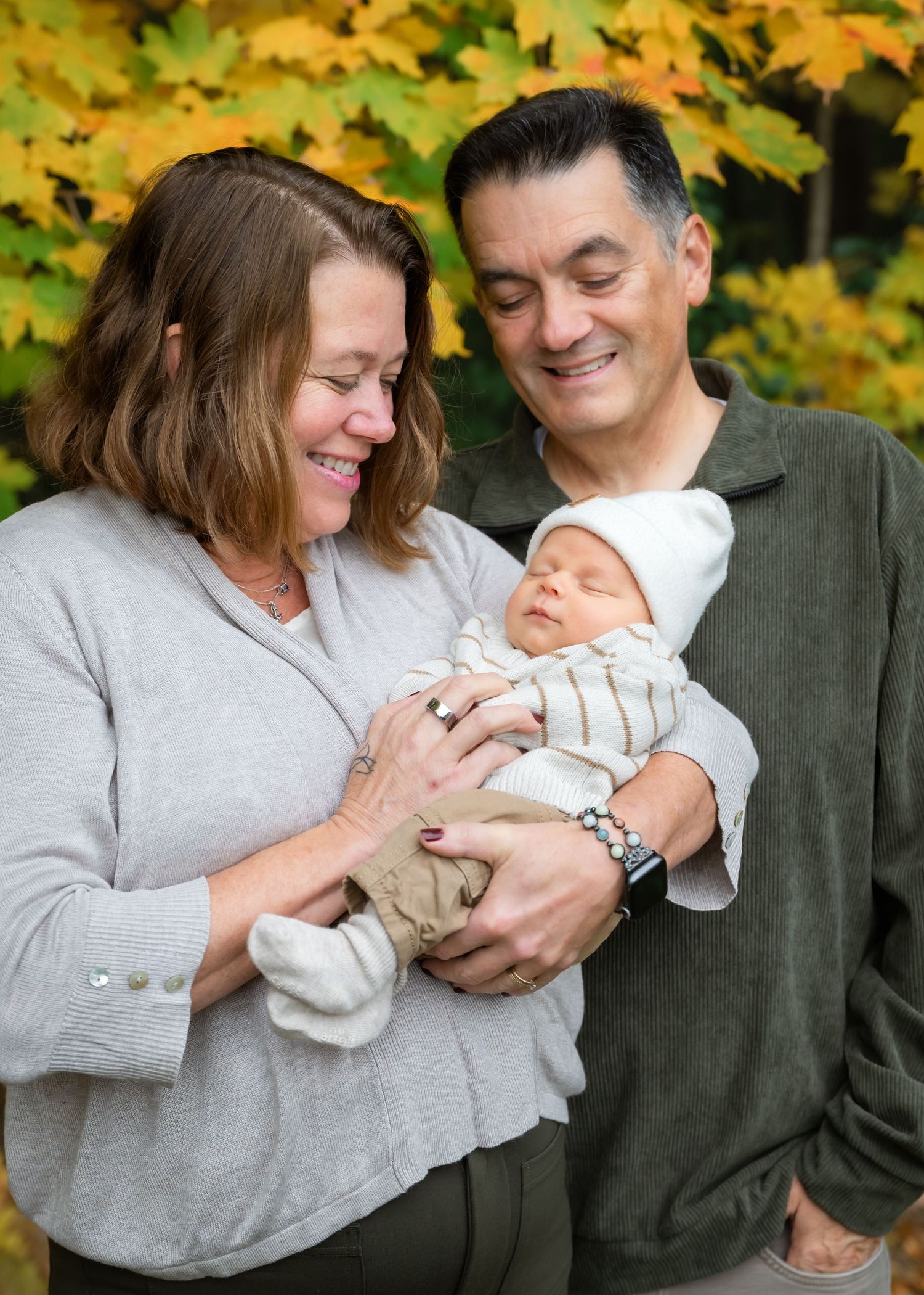 A smiling couple holds a sleeping newborn baby outdoors with vibrant yellow autumn foliage in the background.