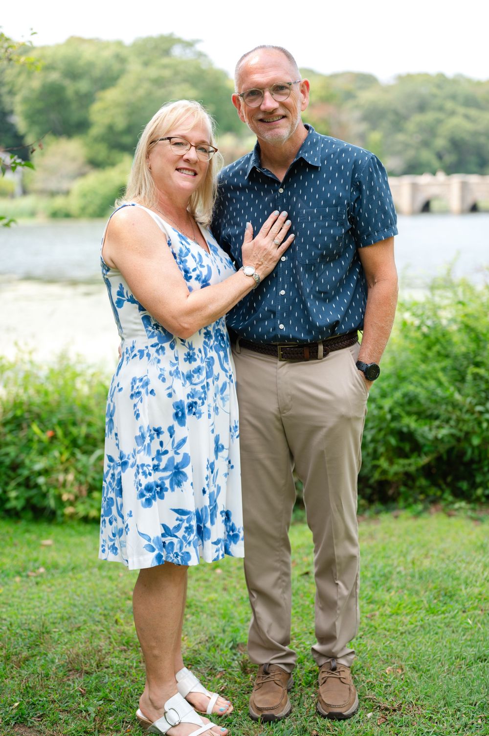 Smiling couple posing outdoors near a river, woman in floral dress and man in navy shirt.