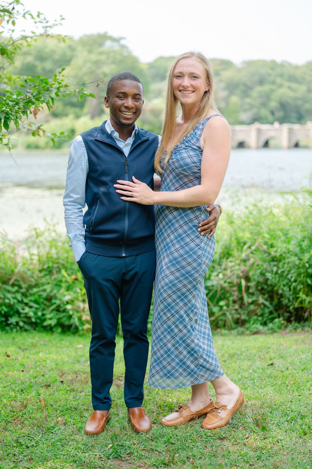 Happy interracial couple posing together outdoors near a river, man in navy vest and woman in plaid midi dress.