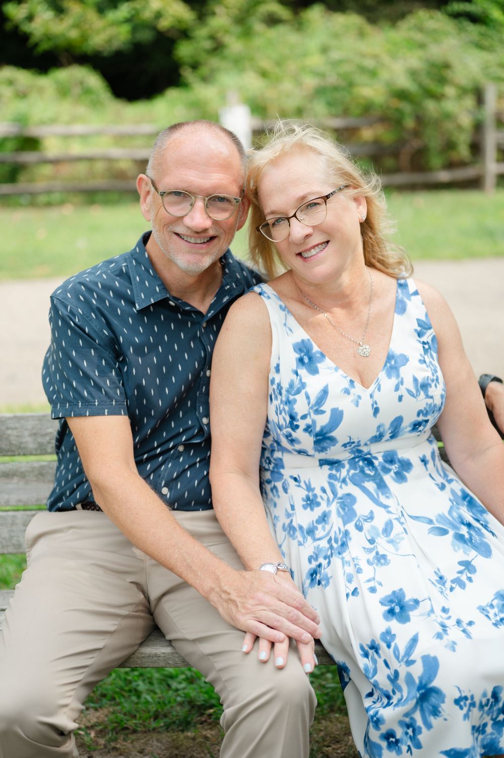 Smiling middle-aged couple sitting on a bench outdoors, man in blue shirt and woman in floral dress, both wearing glasses.