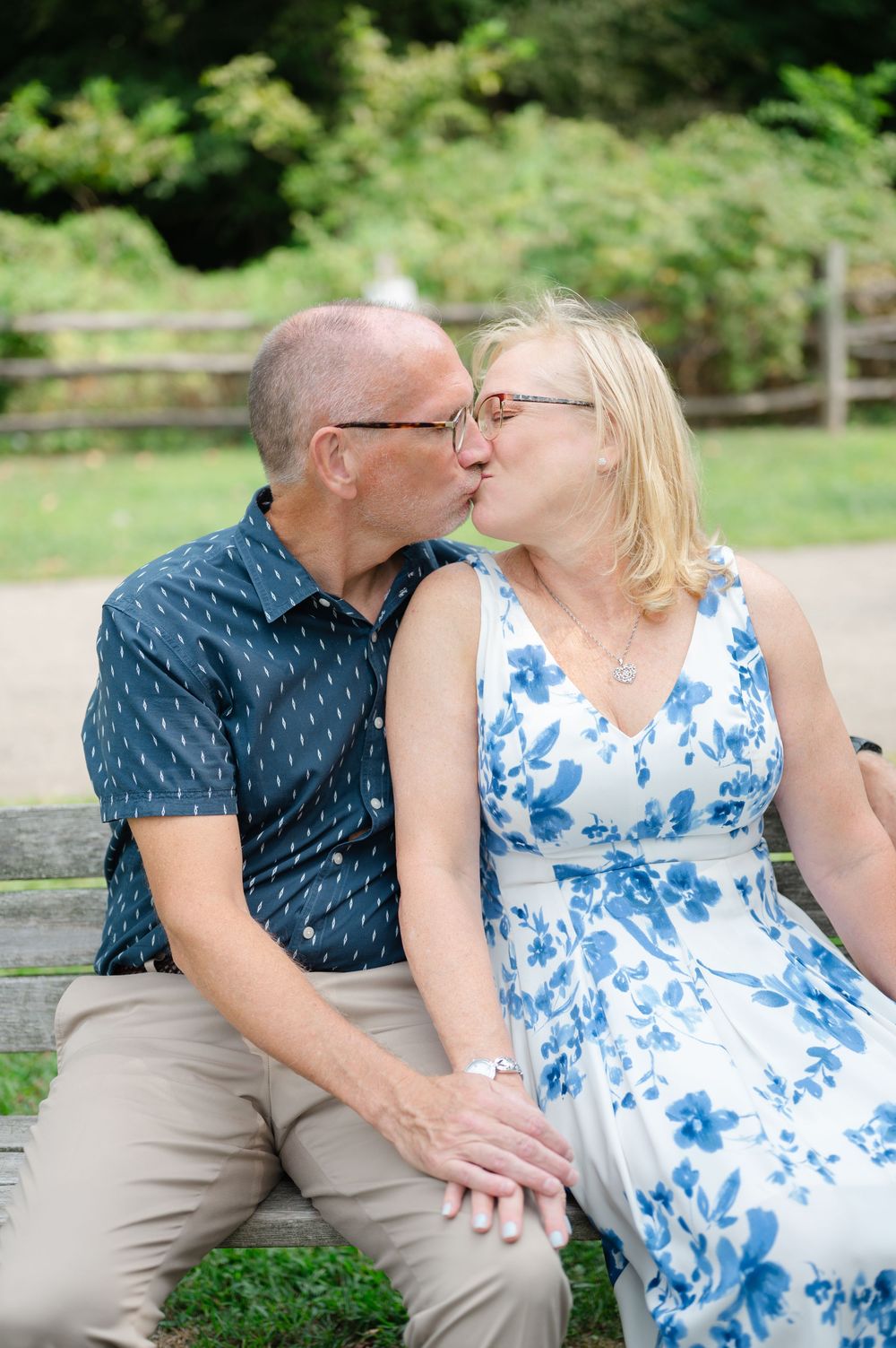 Middle-aged couple sharing a kiss on a park bench, woman in blue floral dress and man in patterned blue shirt.