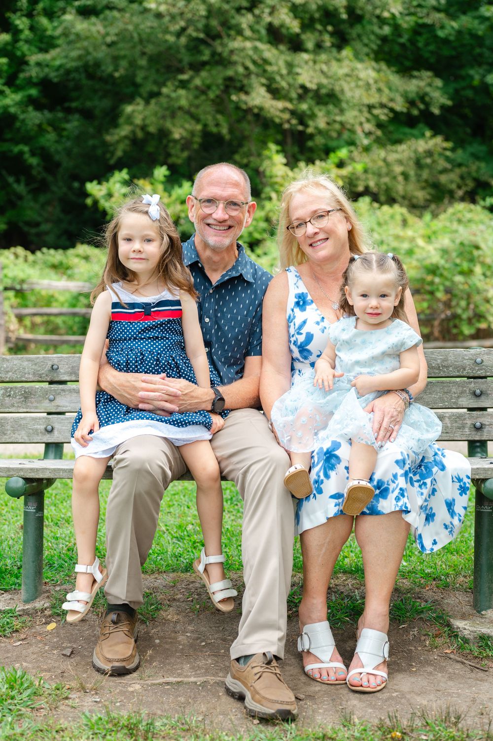 Grandparents sitting on a park bench smiling with two young granddaughters in blue dresses outdoors.