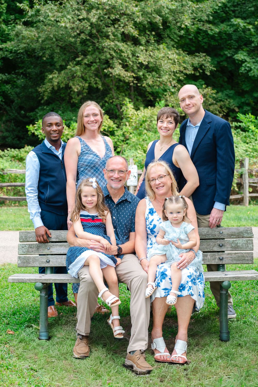Family portrait of seven people posing outdoors on a park bench surrounded by lush green trees.
