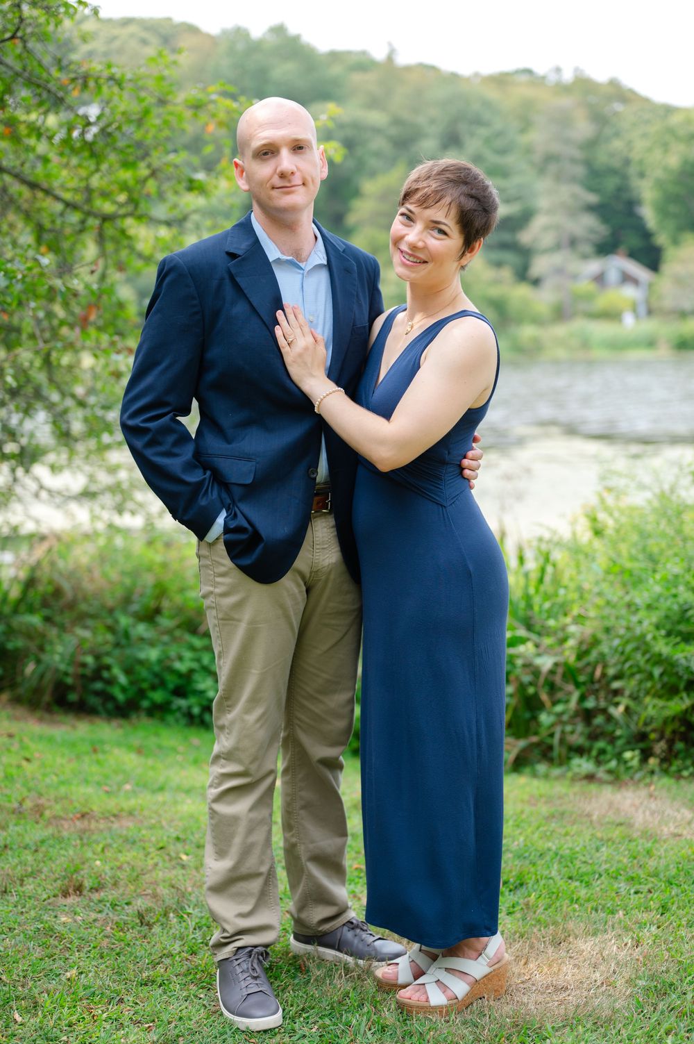 Couple in navy outfits posing outdoors near a lake surrounded by lush green trees.