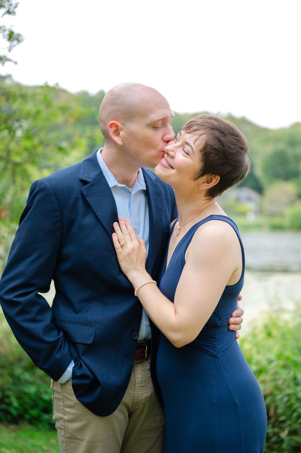 Man in navy blazer kissing woman in blue dress outdoors near a pond.