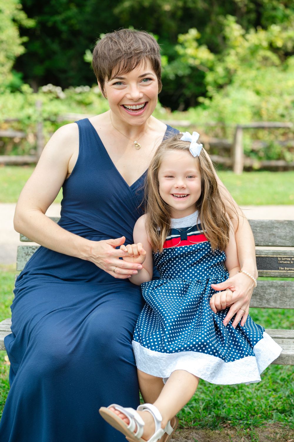 Laughing woman in navy dress sitting on bench with young girl in polka dot dress outdoors.