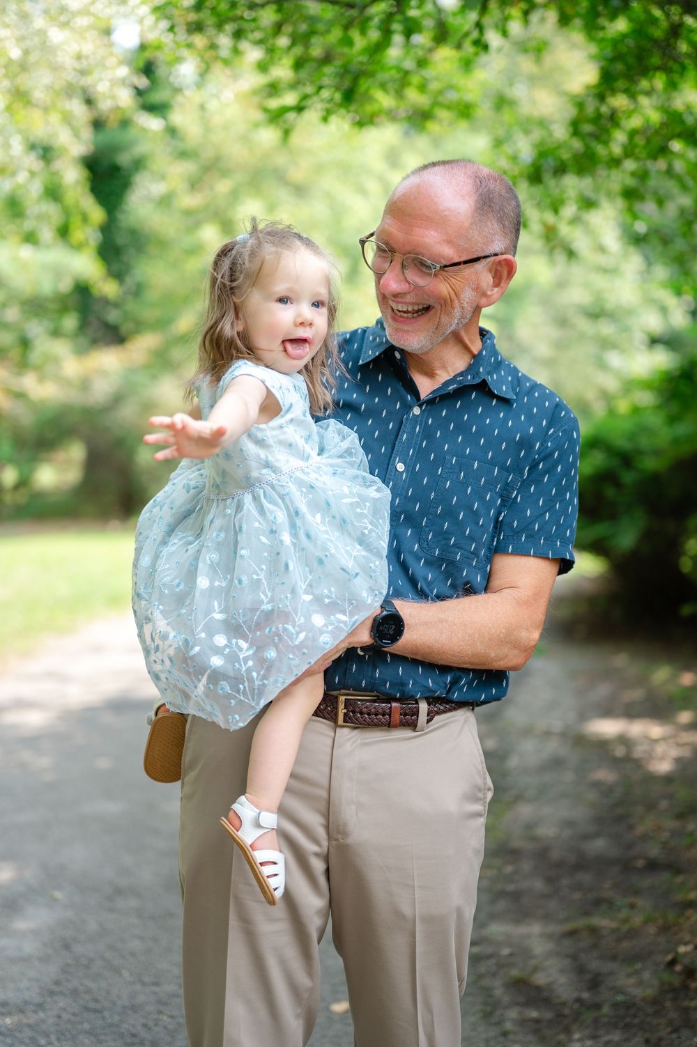 A grandfather with glasses holds a laughing toddler girl in a white dress outdoors on a tree-lined path.