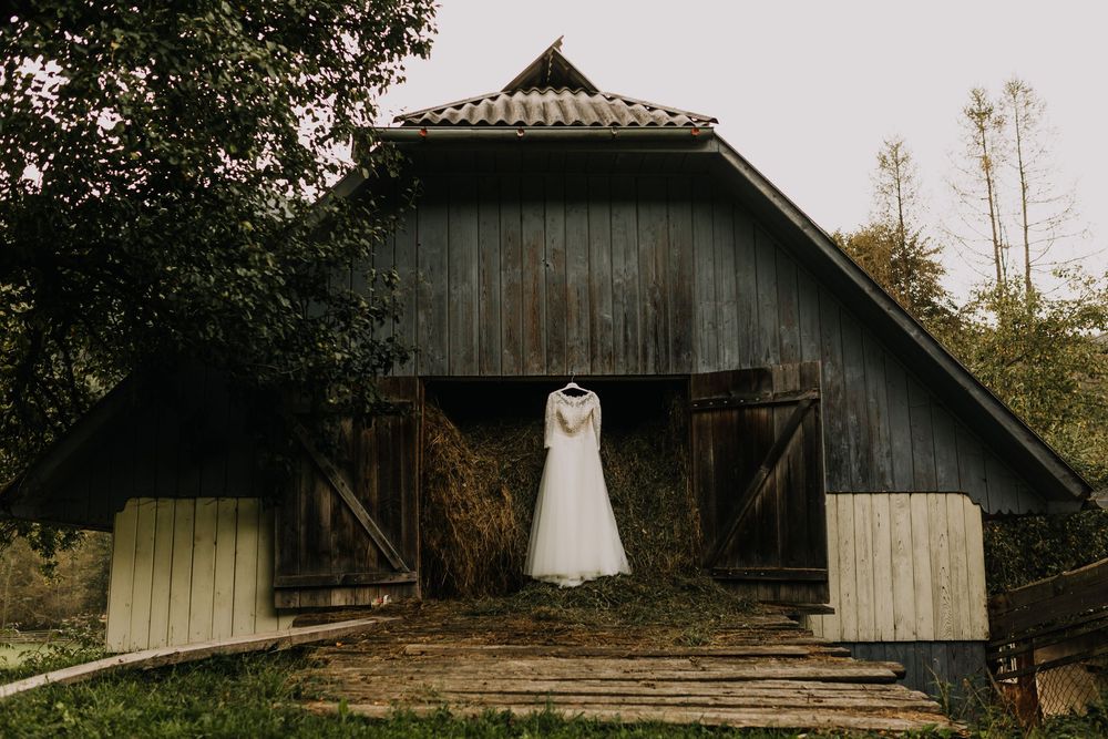 Beautiful white wedding dress hanging on rustic wooden barn doors surrounded by lush green countryside scenery.