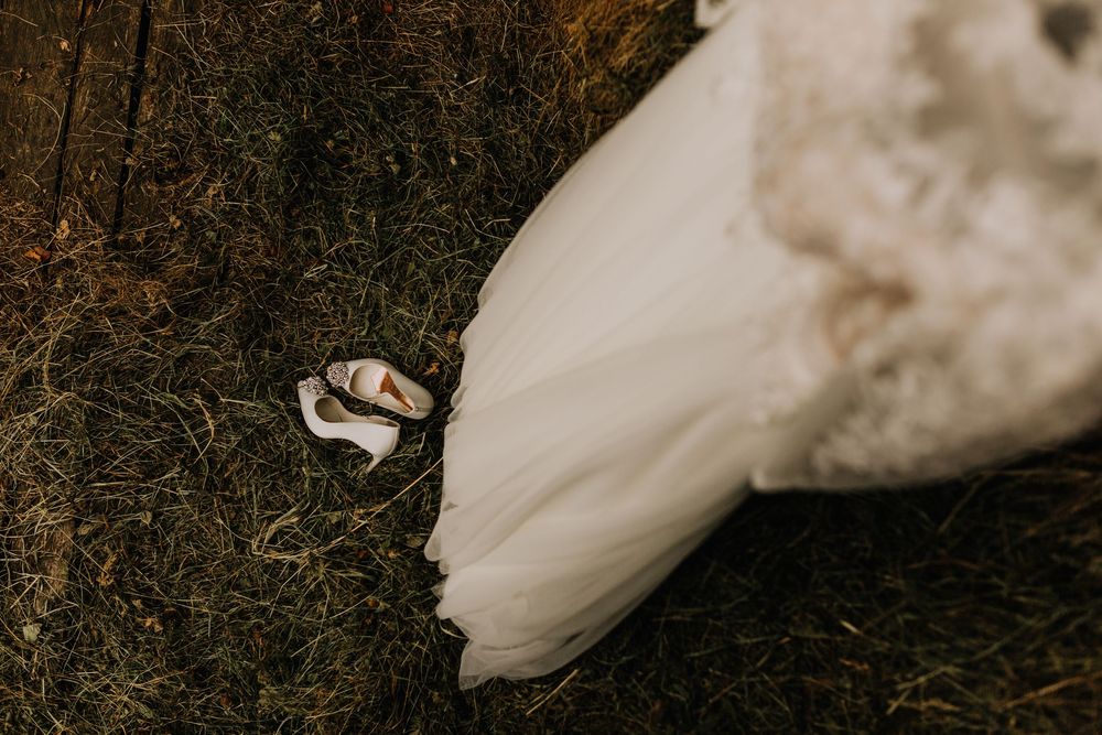 Close-up of bridal gown train spread across hay with delicate white wedding shoes beside it.