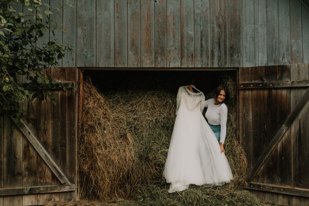 Bride in casual attire carrying elegant white wedding dress through rustic barn filled with golden hay bales.