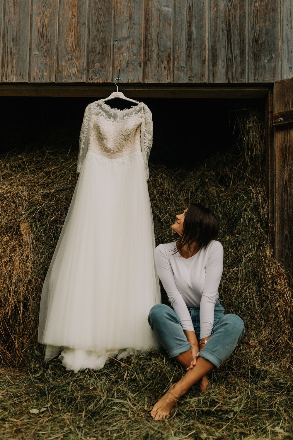 Bride sitting barefoot on hay admiring her lace wedding gown hanging in a rustic wooden barn.