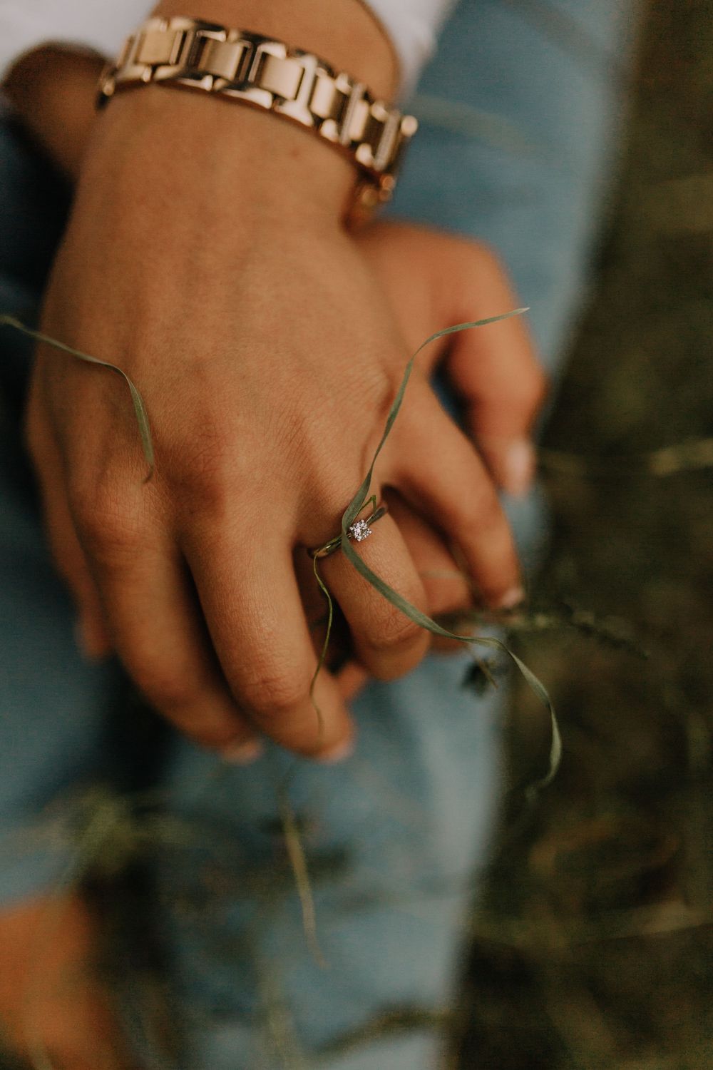 Close-up of bride and groom holding hands, showing engagement ring and gold bracelet in grass.