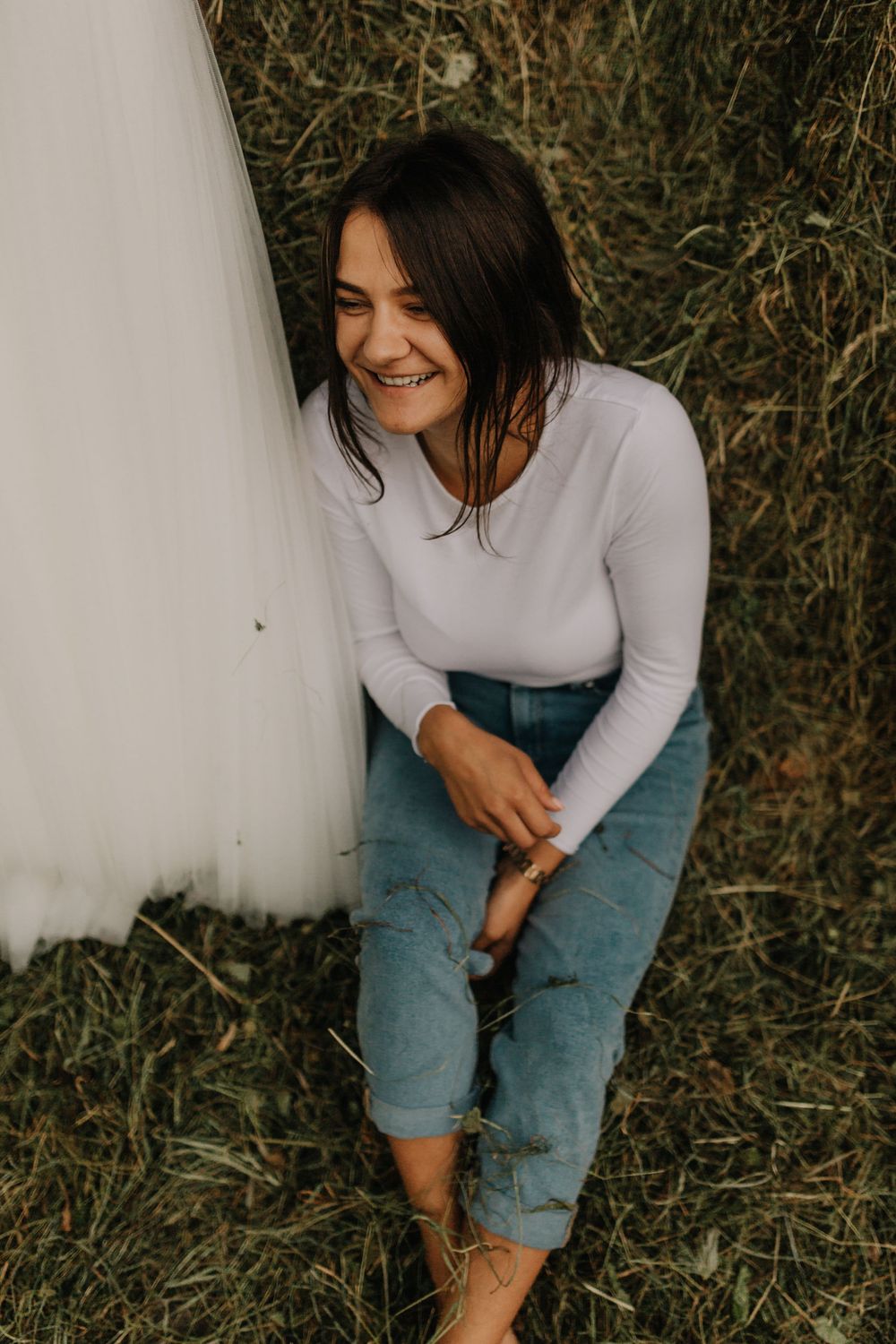 Smiling bride in white top and jeans sitting relaxed in a grassy field beside her wedding dress.