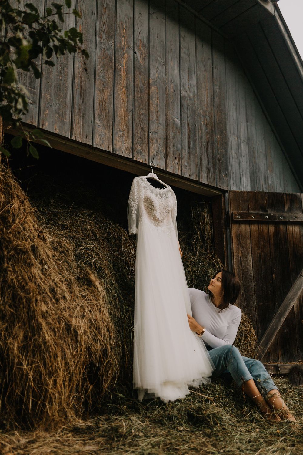 Bride in casual jeans admires her elegant lace wedding gown hanging outside a rustic wooden barn.