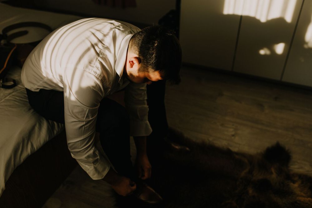 A man in a white shirt bends down to pet a cat on the floor in a dimly lit bedroom.