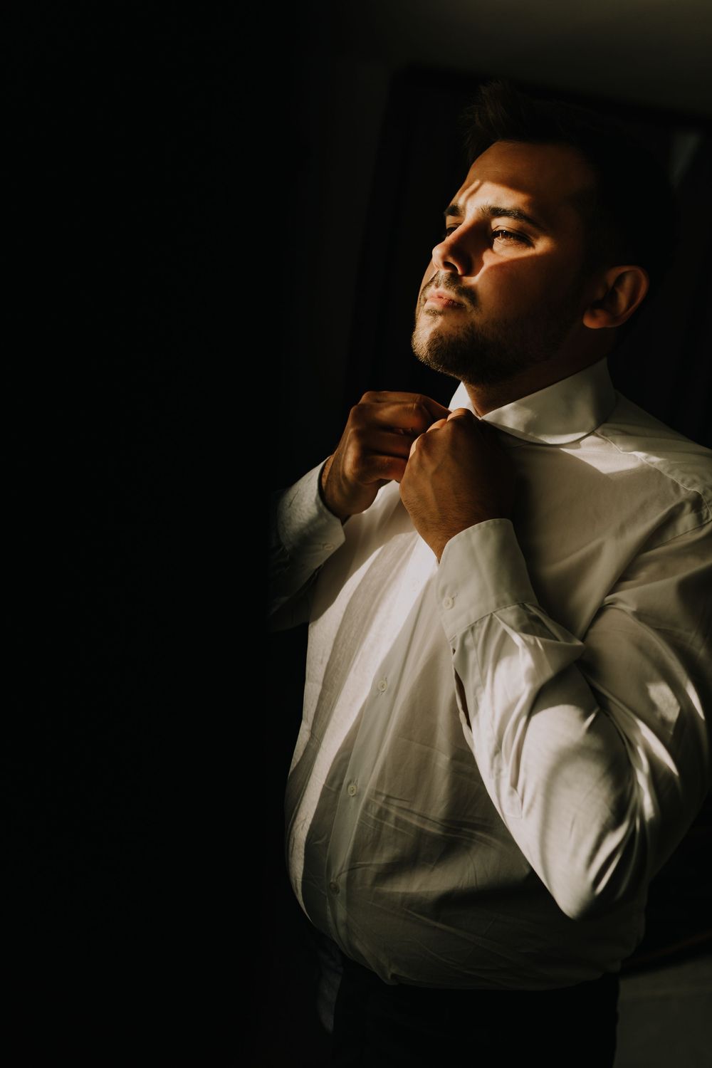 A well-dressed man in a white shirt adjusts his collar in dramatic moody lighting.