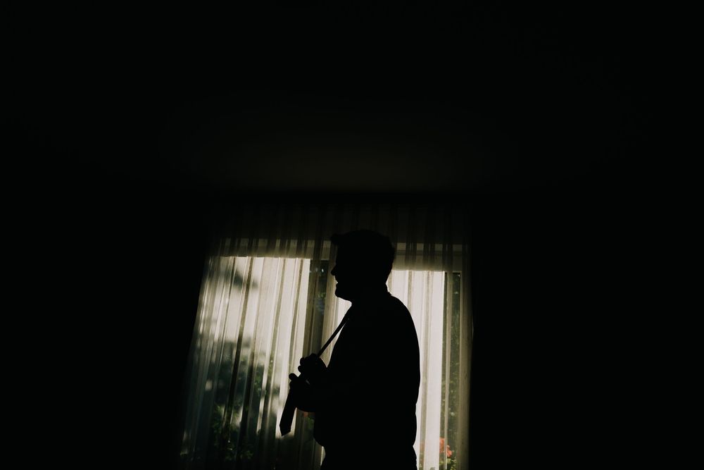 Silhouette of a man standing by a curtained window in a dark room.