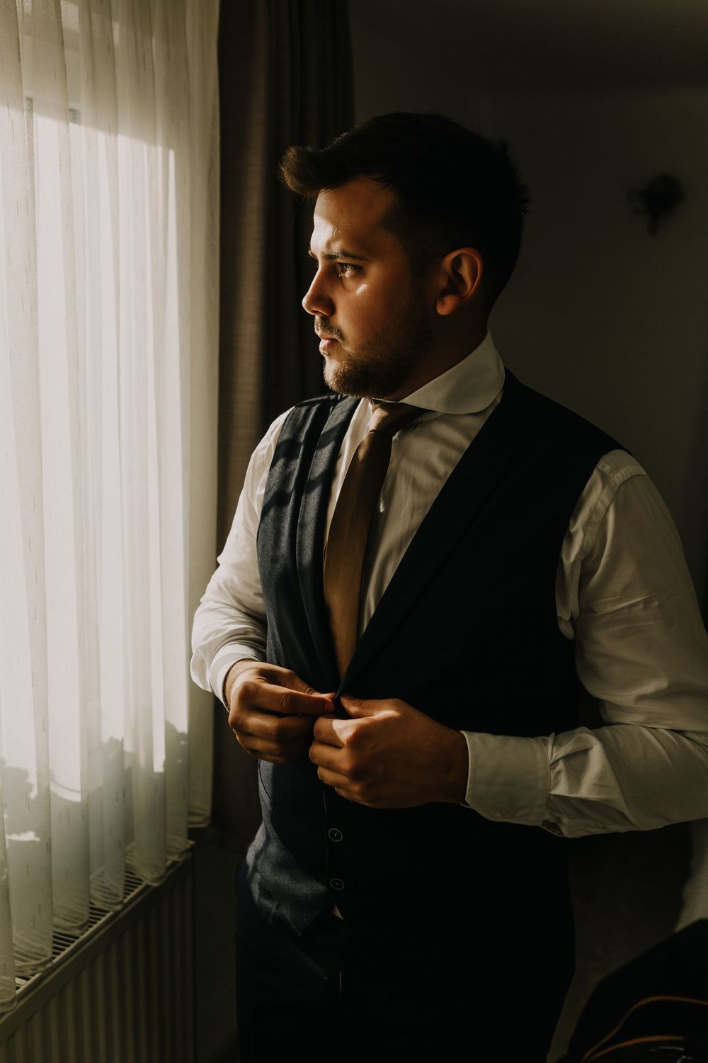 A bearded groom in a vest and tie buttons his waistcoat while gazing out a sunlit window.
