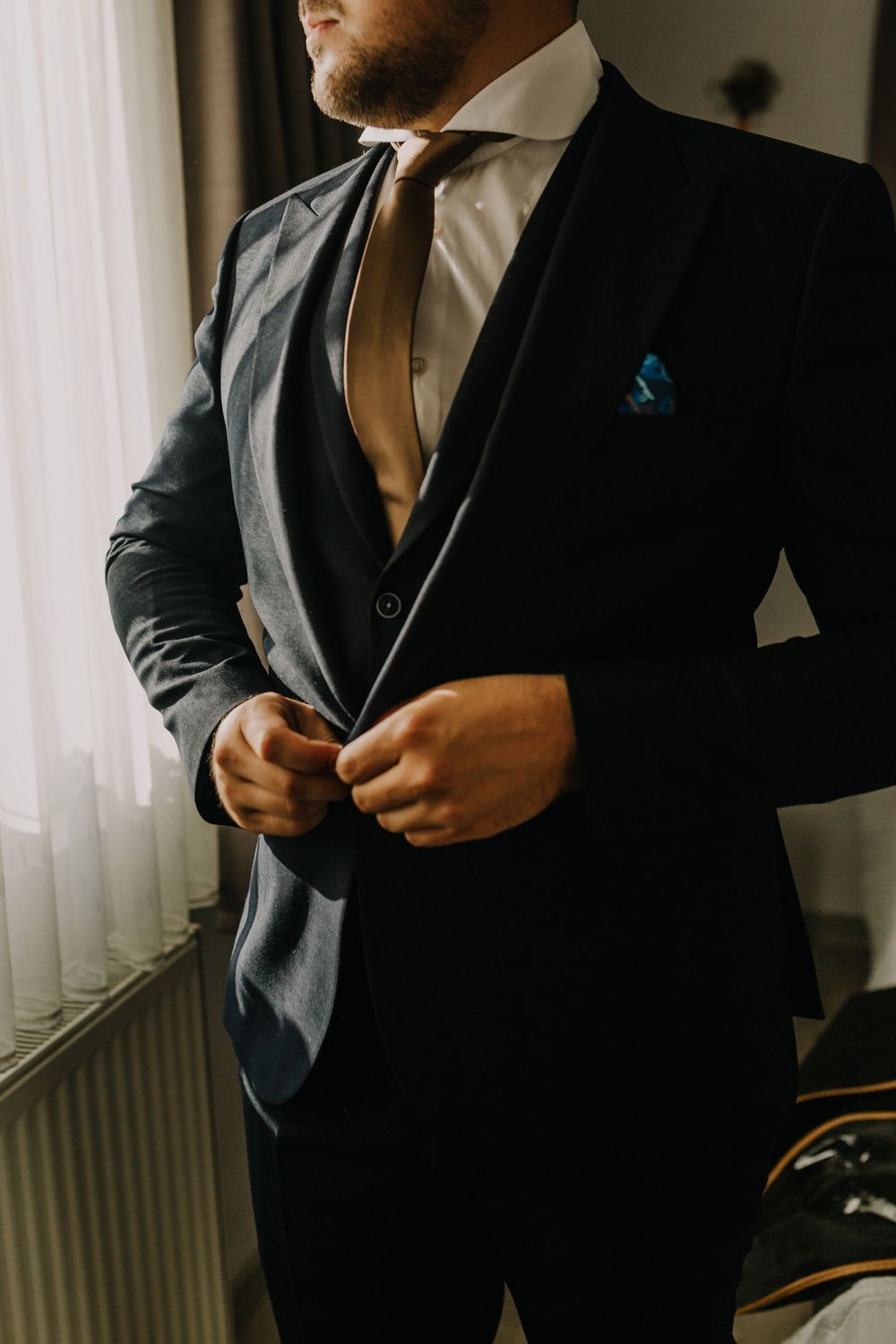 A groom in a dark three-piece suit with gold tie and blue pocket square buttons his jacket.