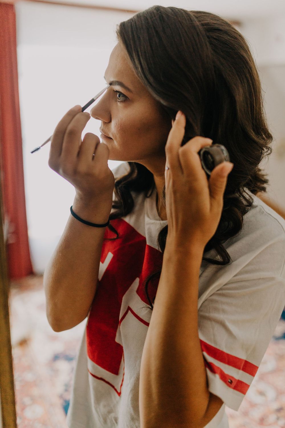 Bride applying eye makeup while looking in a mirror during wedding morning preparations.