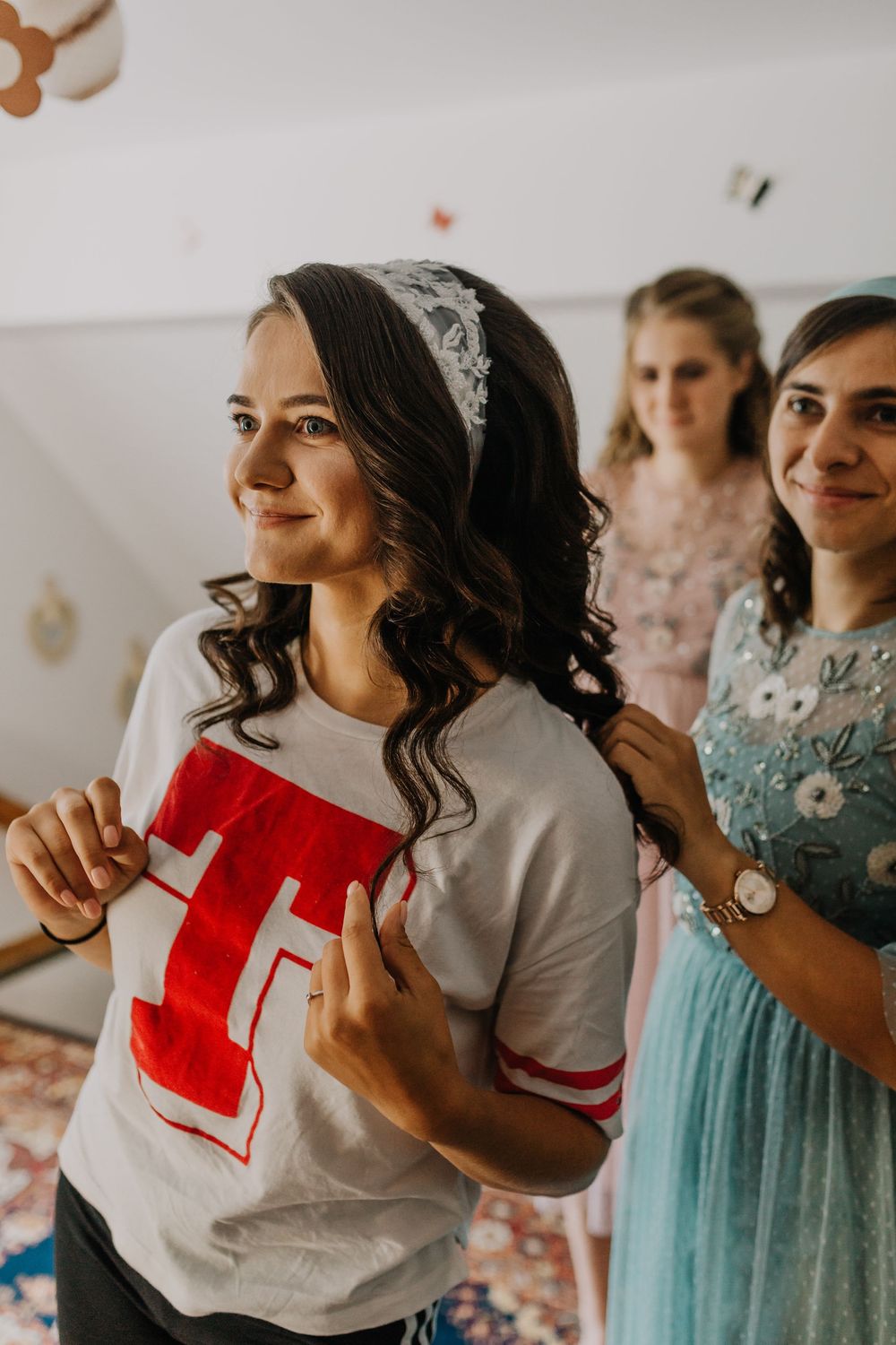 A smiling bride in a white T-shirt gets helped by bridesmaids while wearing a lace headband.